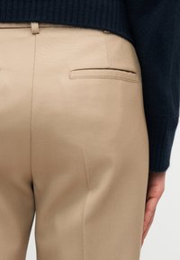 Μπεζ ραμμένο παντελόνι με πίσω τσέπη welt, φορεμένο με σκούρο μπλε πλεκτό πουλόβερ με ρίγες, που δείχνει μέρος του χεριού ενός ατόμου στο πλάι.