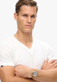 Męska biała koszulka V-neck z teksturowanej tkaniny, noszona z zegarkiem na pasku ze złotej i brązowej skóry, z niebieską tarczą i srebrnymi akcentami.