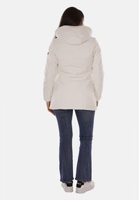 Parka bianca isolata con un ampio cappuccio, caratterizzata da una vita aderente, una texture liscia e senza chiusure visibili, indossata sopra jeans blu.