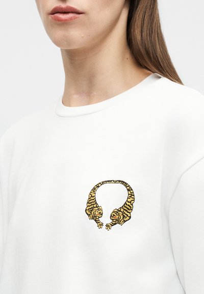 Κοντινή εικόνα ενός ατόμου που φοράει λευκό πουκάμισο με κεντητό σχέδιο δύο κίτρινων τίγρεων που κοιτάζονται μεταξύ τους στο στήθος.