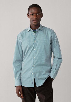 Joven con una camisa de botones a rayas verticales verdes y blancas y pantalones oscuros de pie frente a un fondo claro y liso.