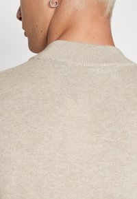 Person mit kurzem blonden Haar und Ohrlochstechen trägt einen beigen, gerippten Rundhalsausschnitt-Pullover, von hinten und im Profil gezeigt.