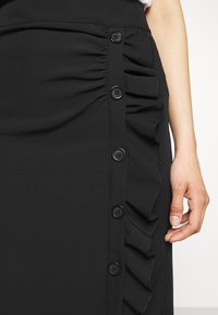 Černá sukně s řasením a volány na straně, se šesti černými knoflíkovými uzávěry viditelnými na levé straně. Hladká textilie.