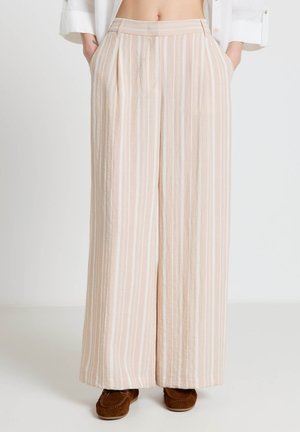 Lower body of person wearing beige and white vertical striped wide-leg trousers with hands in pockets and brown suede shoes.