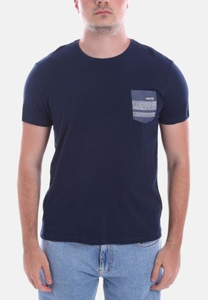 Uomo che indossa una maglietta blu navy con una tasca sul petto con motivo etichettata "YES-ZEE" e jeans azzurri, in piedi davanti a uno sfondo semplice.