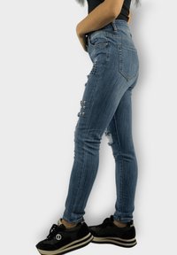 Jeans ajustados de mezclilla con un lavado intermedio, con detalles desgastados y tachuelas plateadas en un lado. Combinados con zapatillas negras con una suela gruesa.