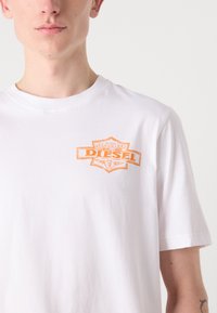 T-shirt bianco in cotone con una patch del logo arancione Diesel sul lato sinistro del petto. Le maniche corte e il collo a giro classico offrono una vestibilità casual.