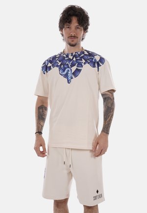 Homme aux cheveux bouclés et tatouages portant un short crème et un T-shirt assorti avec un motif de serpent bleu autour du col.