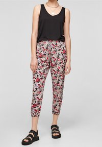 Haut noir sans manches associé à un pantalon clair à motif floral avec des accents rouges, roses et violets. Le mannequin porte des sandales compensées noires.