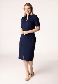 Marineblaues Kleid mit kurzen Ärmeln, Reißverschluss vorne und zwei gesteppte Taschen. Es hat einen knielangen Schnitt und einen Schlitz vorne für zusätzliche Details.