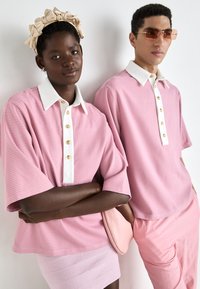 Két személy egymástól függetlenül azonos rózsaszín, rövid ujjú pólóinget visel, amelynek fehér színe és arany gombjai vannak, texturált rózsaszín alsó ruházattal párosítva.