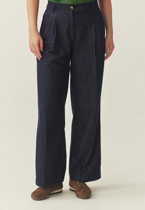 Donna che indossa pantaloni ampi blu scuro, top verde e scarpe stringate marroni, in piedi con le braccia rilassate ai lati.