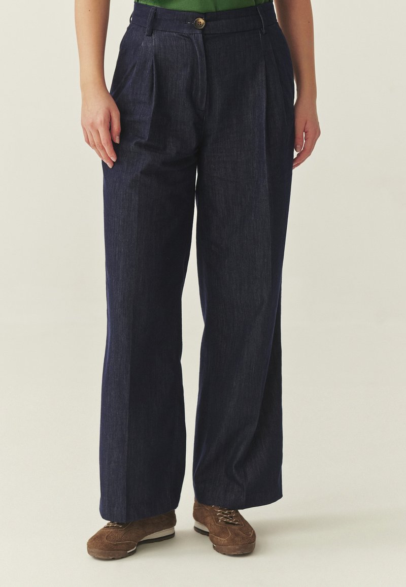 Žena v širokých tmavomodrých nohaviciach, zelenom topu a hnedých šnurovacích topánkach, stojaca s rukami uvoľnene pri tele.