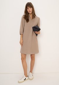 Femme portant une robe beige midi tenant une veste noire pliée, se tenant contre un fond uni clair avec des baskets blanches.