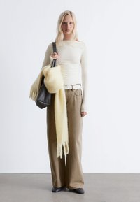 Light cream long-sleeve top, wide tan pants, white belt, carrying a black bag and a fluffy pale yellow scarf with fringe detailing.