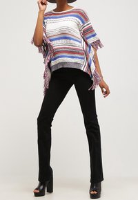Poncho lavorato a maglia multicolore con strisce blu, rosse e crema, dettagli frangiati. Abbinato a pantaloni neri a zampa d'elefante e tacchi a piattaforma.