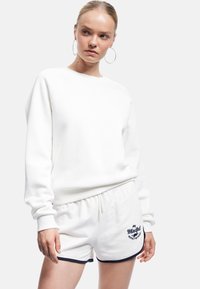 Sudadera blanca de manga larga con puños acanalados, combinada con unos pantalones cortos blancos con ribetes y bordados en azul marino. Textura de tela suave.
