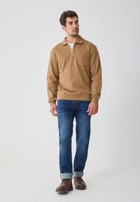 Brun pullover med halv glidelås og teksturert overflate, med brystlomme. Kombinert med blå jeans med oppbrettede ankler og brune støvler.