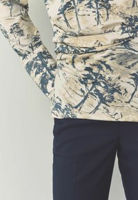 Dlhý rukáv v béžovej farbe s modro-zeleným potlačou stromu, v kombinácii s upravenými námorníckymi nohavicami. Mäkka, textúrovaná tkanina s jemným zberaním v páse.