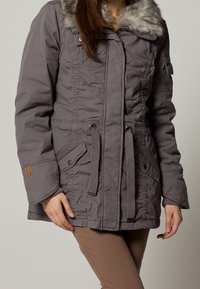 Parka grigia con colletto in pelliccia sintetica, vita regolabile con cordino e chiusura con zip. Ha tasche laterali e tessuto testurizzato.