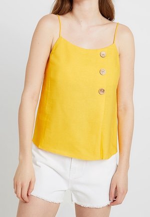 Canottiera gialla in lino con spalline sottili, scollatura dritta e tre bottoni decorativi sul lato sinistro. Abbinata a pantaloni corti bianchi.