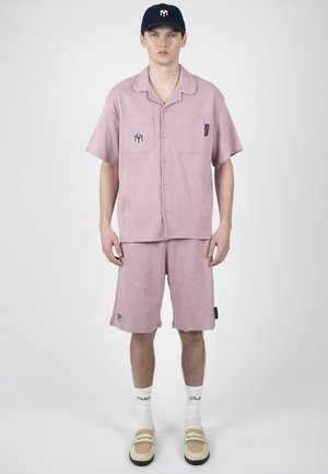 Jeune homme debout portant une chemise à manches courtes rose poussiéreux à boutons et un short assorti, des chaussettes blanches, des chaussures beiges sans lacets et une casquette bleu marine.