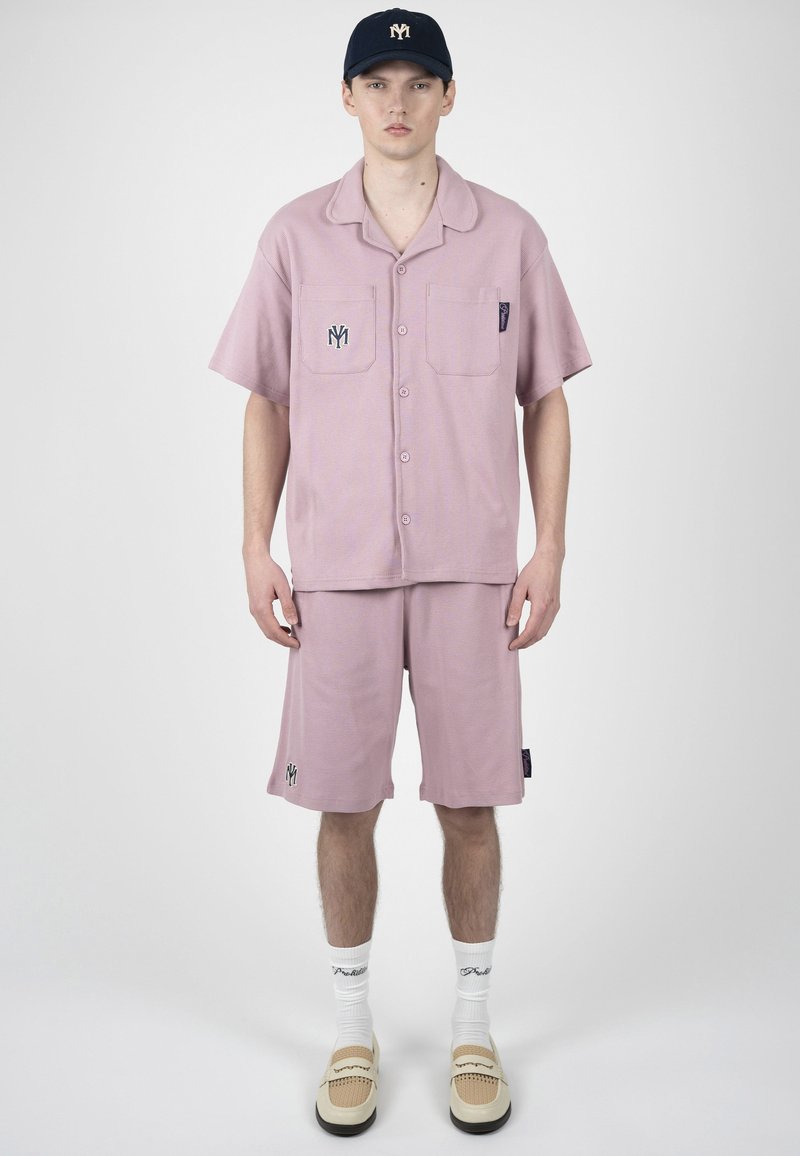 Giovane uomo in piedi che indossa una camicia rosa polveroso a maniche corte con bottoni e pantaloncini abbinati, calzini bianchi, scarpe beige senza lacci e un cappello blu navy.