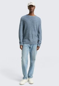 Blauwe gebreide trui met ronde hals, gecombineerd met lichtblauwe jeans en witte sneakers. Een beige pet maakt de casual look compleet.