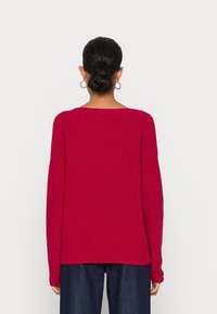 Langere mouwen, geribbelde breisweater in felrood met een ontspannen pasvorm en brede halslijn, gecombineerd met donkere spijkerbroeken, gezien vanuit de achterkant.