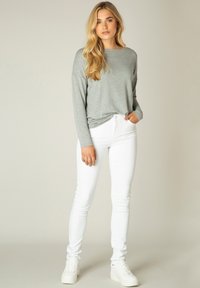 Grijze longsleeve shirt met een relaxte pasvorm, gecombineerd met witte slim-fit jeans. Model draagt witte sneakers en staat tegen een neutrale achtergrond.