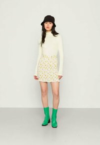 Camisola branca de manga longa, saia mini com padrão floral, botas de tornozelo verdes e um chapéu bucket preto. Design simples com silhueta ajustada.