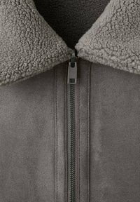 Grijze jas met een getextureerde, pluizige kraag. Voorzak met ritssluiting, afgewerkt met gladde stof en subtiele stikseldetails op het lichaam.