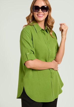 Femme souriante portant des lunettes de soleil foncées et une chemise verte vive à boutons avec les manches retroussées, posant avec une main levée.
