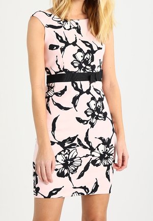 Woman wearing a sleeveless fitted pink dress with large black floral prints and a black waist belt with a bow.