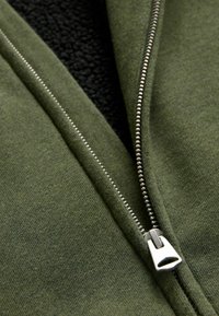 Giacca verde con zip, dotata di un interno in pile testurizzato e un esterno liscio. Presenta una zip metallica lungo l'apertura frontale.