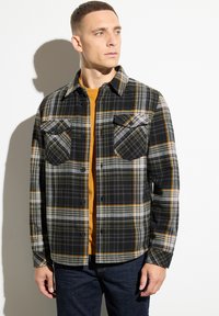 Camicia in flanella a quadri nera e grigia con accenti gialli, dotata di due tasche sul petto, chiusura con bottoni e colletto aperto.