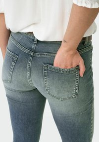 Išblukę mėlyni džinsai su tiesiu kirpimu, turintys dvi galines kišenes ir lygią tekstūrą. Dėvimi su balta palaidine.