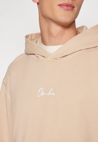 Beige mikina s kapucí a vyšitým logem "Osaka" v bílé barvě, vyrobená z měkké látky, s uvolněným střihem a klokaní kapsou.