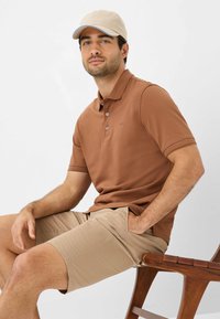 Brun polo skjorte med tekstureret overflade, tre knapper og korte ærmer, kombineret med lyse beige shorts og en beige kasket.