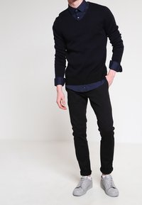 Czarny sweter z dekoltem w serek noszony na ciemnoniebieskiej koszuli z guzikami, w zestawieniu z czarnymi dopasowanymi spodniami i szarymi trampkami. Prosty i nowoczesny design.