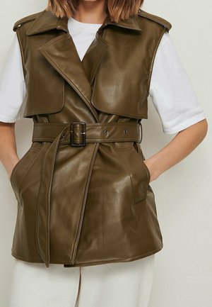 Veste sans manches en cuir vert olive avec larges revers et ceinture, portée sur une chemise blanche à manches courtes, les mains dans les poches.