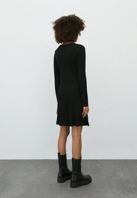 Schwarzes geripptes Strickkleid mit langen Ärmeln und ausgestellt geschnittenem Saum, kombiniert mit klobigen schwarzen Stiefeln. Glatte Textur, tailliertes Design, das den Körper umschmeichelt.
