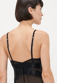 Schwarzes, halbtransparentes Kleid mit dünnen verstellbaren Trägern, einem taillierten Oberteil und geschichteter Textur detailliert entlang des Rückens. Glatte Oberfläche und elegantes Design.
