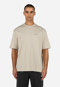 Beige lyhythihainen t-paita, joka on valmistettu pehmeästä kankaasta. Paita on varustettu pyöreällä kaula-aukolla ja vasemmassa rinnassa on minimaalinen brodeerattu logo.