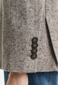Manica di un blazer in tweed marrone con quattro bottoni scuri, che mette in evidenza un tessuto strutturato con piccole screziature, aggiungendo profondità al design neutro.