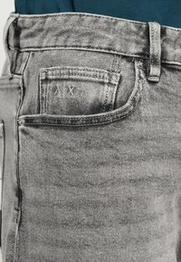 Grå denimjeans med en sydd ficka med initialerna "AX" och ett klassiskt midjeband. Tyget har en lätt blekt textur.