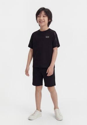 Ragazzo sorridente in piedi, indossa una maglietta nera EA7 e pantaloncini con sneakers bianche su uno sfondo bianco uniforme.