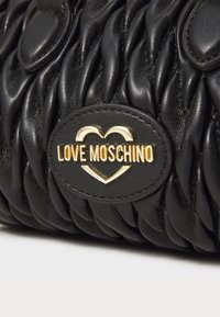Schwarze Lederhandtasche mit einem gesteppte Design, versehen mit einem goldenen Logo-Abzeichen, das eine Herzform enthält. Texturierte Oberfläche mit abgerundeten Akzenten.