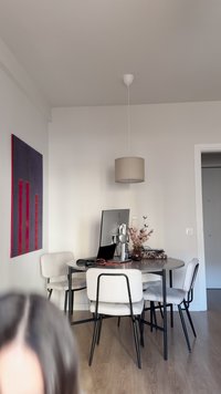 Mesa redonda gris rodeada por cuatro sillas tapizadas blancas con patas negras. Incluye un monitor de escritorio, un jarrón con flores y arte en la pared.