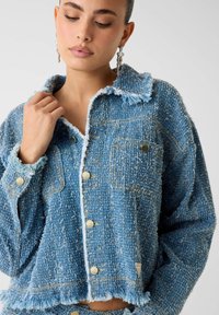 Blauw versleten spijkerjack met gouden draadaccenten, knoopsluiting aan de voorkant, twee borstzakken en een brede kraag, getoond op een model.